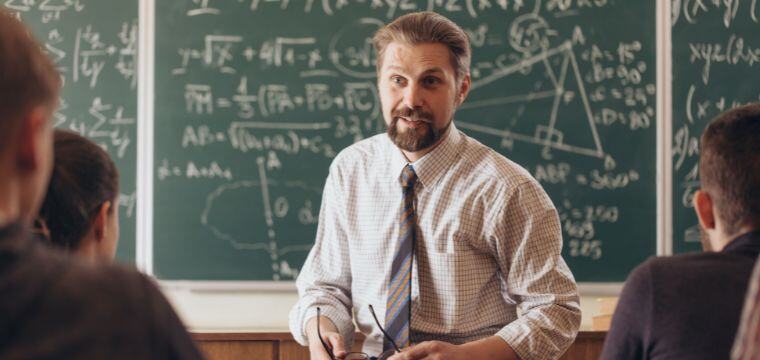 A teacher with a beard stands in front of a chalkboard filled with mathematical formulas, holding glasses, in a classroom setting with students facing him.