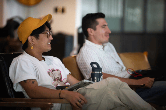 Two people sitting indoors on chairs, one wearing a yellow cap and glasses, both smiling and appearing engaged in conversation or a meeting.