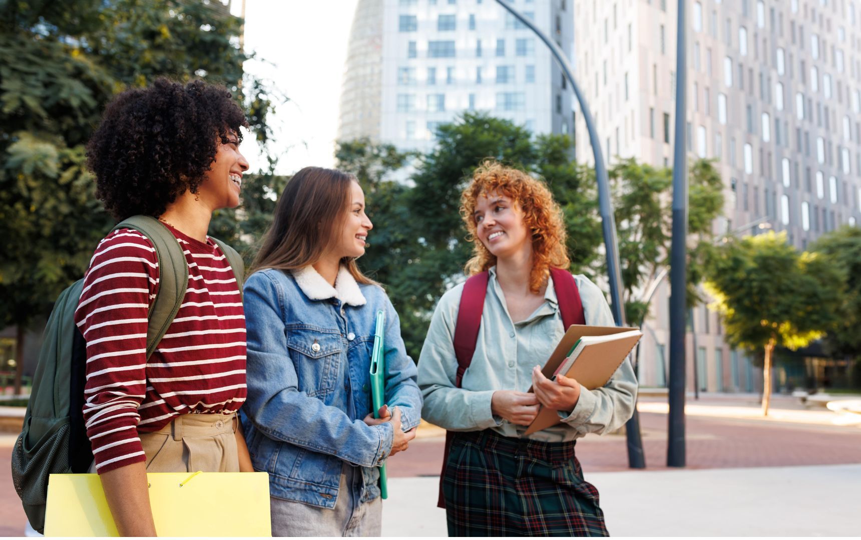 Three young people with backpacks and notebooks are walking and talking together outdoors in an urban area with tall buildings and trees.